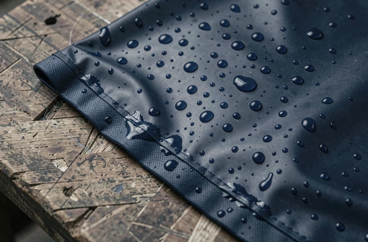 Gros plan sur un tissu enduit imperméable marine avec des gouttes d'eau perlant sur la surface, révélant les limites d'imperméabilité au niveau des coutures
